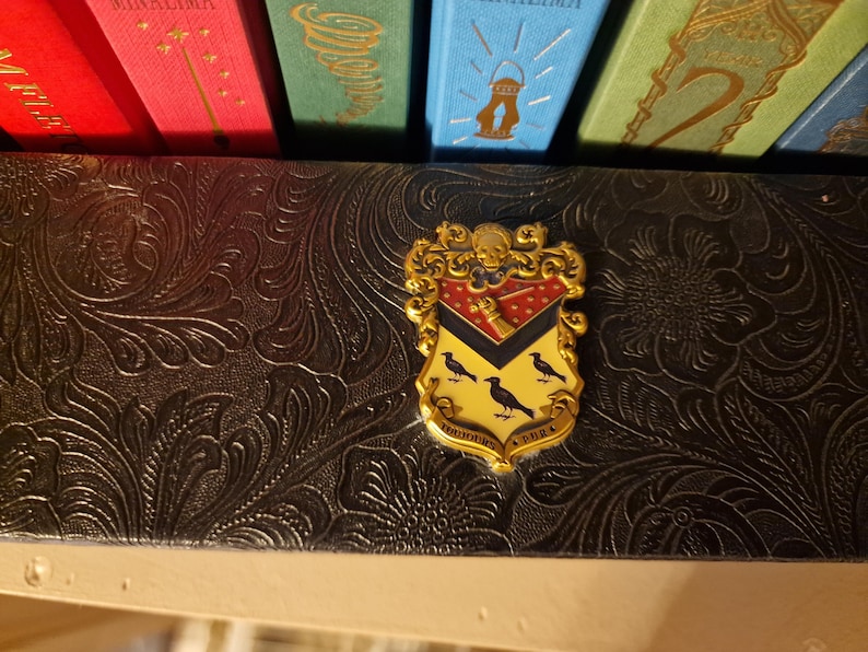 May include: A gold-colored crest pin with a red, black, and yellow shield design, featuring three black birds. The pin is set against a dark, textured background, with colorful book spines visible above.