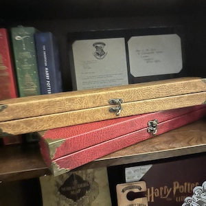 May include: A set of two red and brown leather-bound boxes with metal clasps. The boxes are stacked on top of each other. The boxes are on a wooden shelf with other Harry Potter themed items, including a Marauder's Map, a Hogwarts sign, and a Harry Potter book.