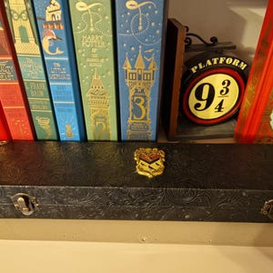 May include: A black, embossed case with gold-colored corner accents and a decorative emblem. The case is positioned in front of a collection of colorful books, including a Harry Potter book and a Platform 9 3/4 sign.