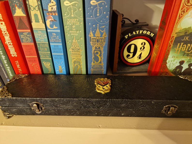 May include: A black, embossed case with gold-colored corner accents and a decorative emblem. The case is positioned in front of a collection of colorful books, including a Harry Potter book and a Platform 9 3/4 sign.
