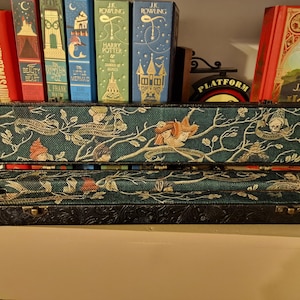 May include: An open, rectangular box with a teal fabric interior featuring a whimsical design of figures, branches, and text. The box has a black, embossed exterior and is displayed with several books in the background.