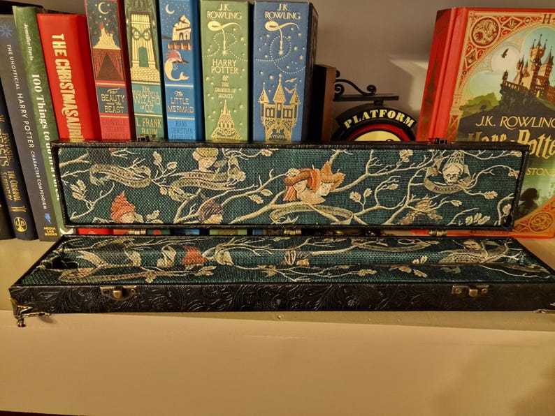 May include: An open, rectangular box with a teal fabric interior featuring a whimsical design of figures, branches, and text. The box has a black, embossed exterior and is displayed with several books in the background.