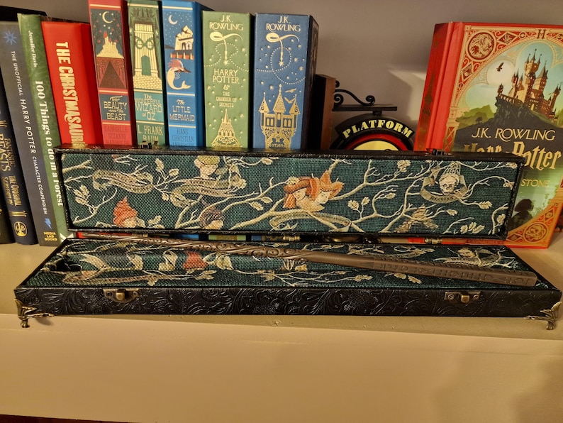 May include: A decorative wand in a black case with a teal and floral design. The case is open, revealing the wand inside. Bookshelves with various books are in the background, including a red book with the title "The Christmasaurus".