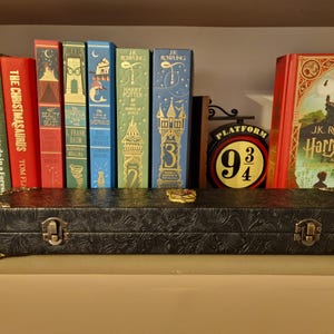 May include: A black, embossed case with metal clasps and decorative corners sits on a shelf in front of a collection of colorful books. The books include titles like "Harry Potter" and "The Christmasaurus". A "Platform 9 3/4" sign is also visible.