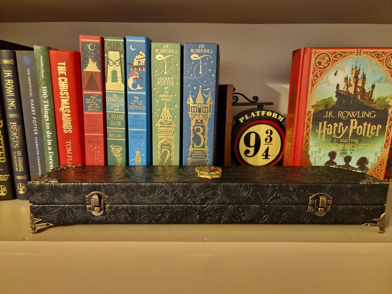 May include: A black, embossed case with metal clasps and decorative corners sits on a shelf in front of a collection of colorful books. The books include titles like "Harry Potter" and "The Christmasaurus". A "Platform 9 3/4" sign is also visible.