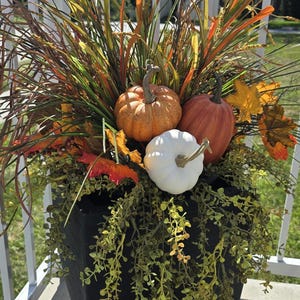 May include: A black square planter overflowing with fall foliage. The arrangement includes orange and white pumpkins, colorful leaves, and various grasses. The planter is on a porch with a white railing.
