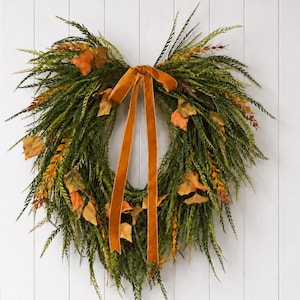 May include: A heart-shaped wreath constructed from dense green foliage and interspersed with orange and yellow autumn leaves. A long, smooth, burnt orange velvet ribbon is tied in a bow at the top center, with two long tails hanging down the middle.