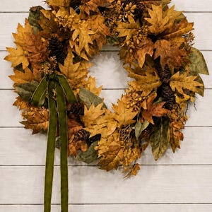 May include: A circular wreath featuring a dense arrangement of dried oak leaves in shades of brown, gold, and muted green. The wreath is accented with dark brown pinecones and small clusters of berries, finished with a long, hanging olive green ribbon.