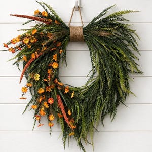 May include: A heart-shaped wreath constructed from dark green fern-like foliage and vibrant orange wildflowers. The arrangement is bound at the top with a thick jute twine loop, which is suspended from a white plastic hook on a paneled wall.