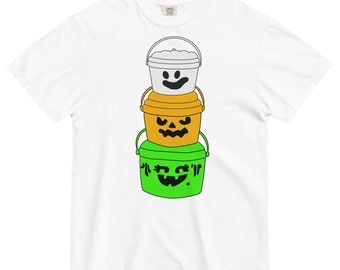 May include: A white t-shirt featuring a graphic of three stacked buckets. The top bucket is white with a smiling face, the middle bucket is orange with a jack-o'-lantern face, and the bottom bucket is green with a smiling face.