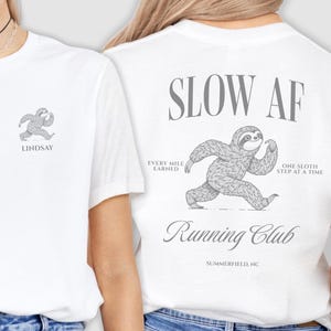 May include: White t-shirt with a running sloth graphic. The front of the shirt has a small sloth graphic and the name "LINDSAY". The back of the shirt has the text "SLOW AF Running Club" with a larger sloth graphic.