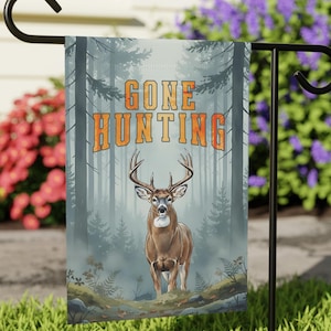 May include: A garden flag with the words "GONE HUNTING" in orange letters. The flag features a detailed illustration of a deer in a forest setting. The background shows tall trees and a misty atmosphere.