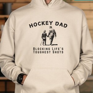 May include: A beige hoodie with a black graphic of a father and son playing hockey. The text "Hockey Dad" is above the graphic and "Blocking Life's Toughest Shots" is below the graphic.