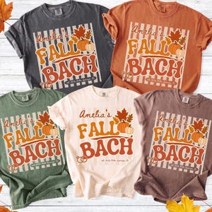 May include: Five short-sleeved crew neck t-shirts in charcoal, burnt orange, sage green, cream, and mauve. Each shirt features a graphic with the text "Amelia's FALL BACH" in orange, accompanied by illustrations of pumpkins, autumn leaves, and rings.