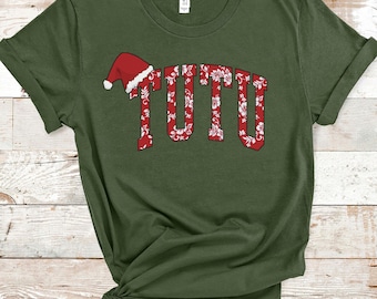 Op de afbeelding: Olijfgroen T-shirt met het woord "TUTU" in een rood, bloemig ontwerp. Een rode kerstmuts staat boven de eerste letter. Het shirt is gemaakt van een zacht materiaal en heeft opgerolde mouwen.
