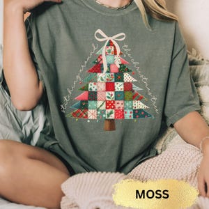 May include: Moss green t-shirt featuring a Christmas tree design made of patchwork squares in red, green, and white. The words "stitching around the Christmas tree" are written around the tree. A white bow is at the top of the tree.