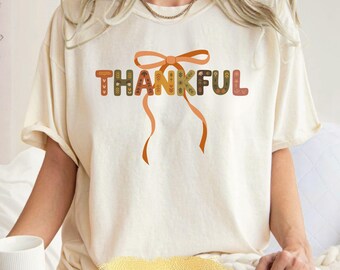 May include: Ivory-colored t-shirt with the word "THANKFUL" in a decorative, fall-themed font. The letters are adorned with patterns in shades of brown, green, and orange, with a bow and ribbons accentuating the design. The shirt is a relaxed fit.
