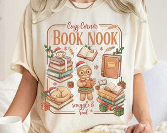 May include: Ivory-colored t-shirt with a graphic that reads "Cozy Corner Book Nook." The design features stacks of books, a cat, a gingerbread person, and other festive elements. The word "snuggle & read" is also included.