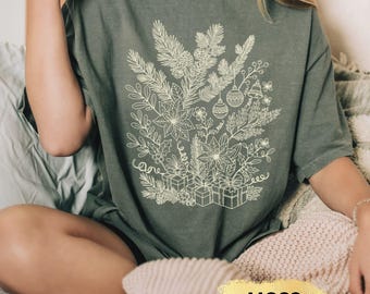 May include: Moss green t-shirt featuring a detailed white floral design with Christmas ornaments, bells, and gift boxes. The shirt has a relaxed fit and is made of a soft material. The word "MOSS" is printed on a gold label.