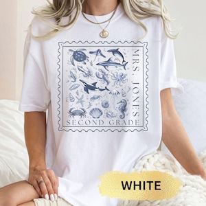 May include: White t-shirt with a blue and white graphic of various sea creatures, including a turtle, dolphins, and seahorse, within a stamp-like border. The text "MRS. JONES SECOND GRADE" is printed on the shirt.