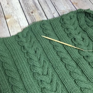 Op de afbeelding: Close-up van een groen gebreid kledingstuk met een kabelbreipatroon. Een houten breinaald is in de stof gestoken. Het kledingstuk ligt op een houten oppervlak.