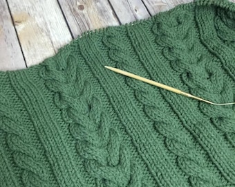 May include: Close-up of a green knitted garment with a cable knit design. A wooden knitting needle is inserted into the fabric. The garment is on a wooden surface.