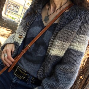 May include: A grey, cream, and light grey striped knitted cardigan with wooden buttons. The cardigan is worn over a grey t-shirt with a peace sign graphic. A brown leather crossbody bag and a black studded belt complete the outfit.