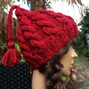 May include: A vibrant red knit hat with a cable-knit design and a long, braided tassel. The hat features a pointed top and a matching tassel at the end of the braid. The hat is made of a soft, textured yarn.