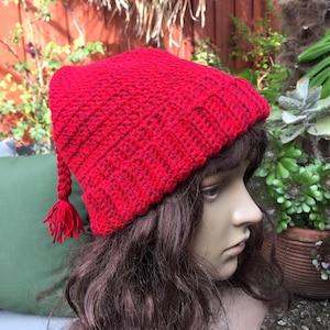 Puede incluir: Un gorro de ganchillo rojo vibrante con una borla. El gorro es de color liso y parece estar hecho de un hilo suave. El gorro es de estilo gorro con una parte superior puntiaguda.