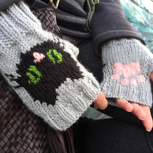 Könnte beinhalten: Ein Paar graue Fingerlose Handschuhe mit gestickten Pfotenabdrücken. Ein Handschuh hat eine schwarze Katze mit grünen und rosa Blumen bestickt.
