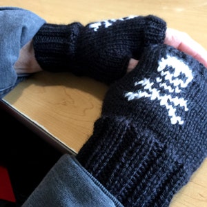 Puede incluir: Guantes sin dedos de punto negro con un diseño de calavera blanca. Los guantes están hechos de un hilo grueso y texturizado y tienen un puño acanalado. El diseño de la calavera está en el dorso de la mano.