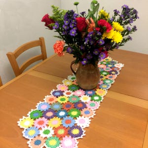 Könnte beinhalten: Ein bunter gehäkelter Tischläufer mit Blumenmuster. Der Läufer ist aus weißem, rosa, gelbem, grünem, blauem und violettem Garn gefertigt. Er liegt auf einem Holztisch mit einer Blumenvase in der Mitte.