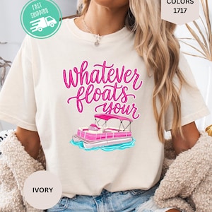 Op de afbeelding: Ivoorkleurig T-shirt met de roze tekst "Whatever floats your" en een roze en blauwe pontonboot-afbeelding. Het shirt maakt deel uit van de Comfort Colors 1717 collectie.