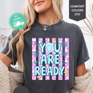 Könnte beinhalten: Dunkelgraues T-Shirt mit den Worten "YOU ARE READY" in Hellblau, einem lächelnden Gesicht und Sternen. Das Shirt hat einen gestreiften Hintergrund mit rosa und weißen vertikalen Linien. Das Shirt ist Teil der Comfort Colors 1717 Kollektion.