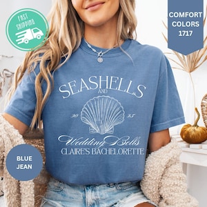 May include: Blue jean t-shirt with the text "Seashells and Wedding Bells Claire's Bachelorette" and a seashell graphic. The shirt is a comfort color and has a round neck.