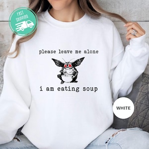 May include: White sweatshirt with a graphic of a creature with large ears and red eyes, holding a bowl. The text on the sweatshirt reads "please leave me alone" and "i am eating soup". A white circle with the word "WHITE" is on the bottom right.
