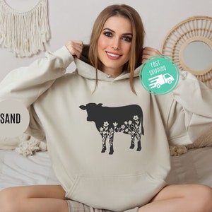 K&ouml;nnte beinhalten: Ein sandfarbener Hoodie mit einer schwarzen Silhouette einer Kuh, aus deren K&ouml;rper Blumen wachsen. Der Hoodie hat eine Kapuze mit Kordelzug und eine K&auml;ngurutasche.