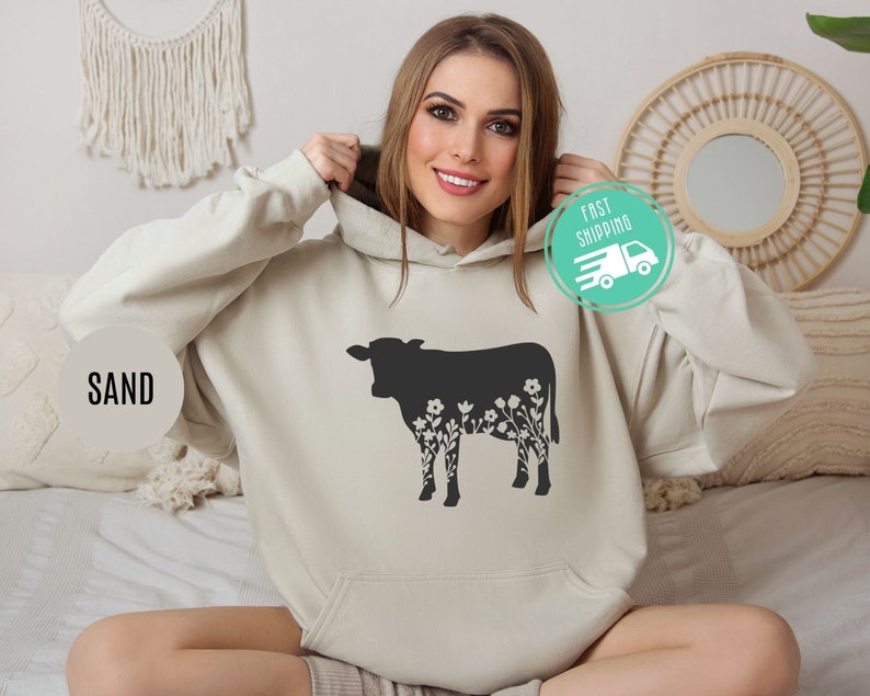 K&ouml;nnte beinhalten: Ein sandfarbener Hoodie mit einer schwarzen Silhouette einer Kuh, aus deren K&ouml;rper Blumen wachsen. Der Hoodie hat eine Kapuze mit Kordelzug und eine K&auml;ngurutasche.