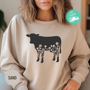 K&ouml;nnte beinhalten: Ein beiger Pullover mit einer schwarzen Silhouette einer Kuh mit floralen Details. Das Wort "SAND" ist in der linken unteren Ecke des Pullovers aufgedruckt.