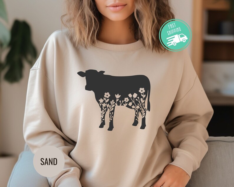 K&ouml;nnte beinhalten: Ein beiger Pullover mit einer schwarzen Silhouette einer Kuh mit floralen Details. Das Wort "SAND" ist in der linken unteren Ecke des Pullovers aufgedruckt.