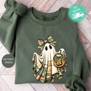 Op de afbeelding: Een militair groene sweater met ronde hals en een centrale afbeelding van een geest met een camouflage strik die een klein oranje pompoenemmertje vasthoudt. De geest is gedrapeerd in een laken met camouflagepatronen aan de onderzoom.