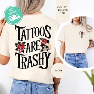 May include: An ivory t-shirt featuring a large black graphic on the back that reads Tattoos Are Trashy, decorated with red roses and a skull. The front chest area displays a smaller matching graphic of a hand holding a red rose with the same text.