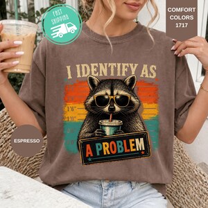 May include: A brown short-sleeve t-shirt featuring a graphic of a raccoon wearing sunglasses and holding a drink. The text reads I IDENTIFY AS A PROBLEM. The background of the graphic includes a height chart and horizontal stripes in orange, yellow, and teal colors.