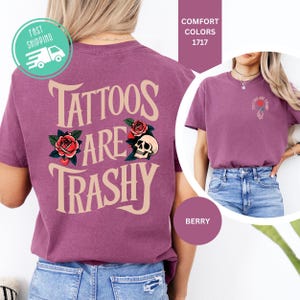May include: A berry-colored t-shirt featuring the text TATTOOS ARE TRASHY in large beige letters on the back, accompanied by red rose and skull graphics. The front chest area displays a smaller version of the same text and a rose graphic.