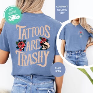 May include: A blue jean colored t-shirt featuring the text "TATTOOS ARE TRASHY" in a large, curved font on the back, accompanied by illustrations of red roses and a skull. The front chest area displays a smaller version of the same graphic.