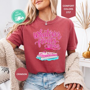 Op de afbeelding: Karmizijnrood T-shirt met de tekst "Whatever floats your" in roze script, met een roze en witte pontonboot afbeelding. Het model draagt een spijkerbroekshort en een cr&egrave;me vest. Een snelle verzendicoon is zichtbaar.