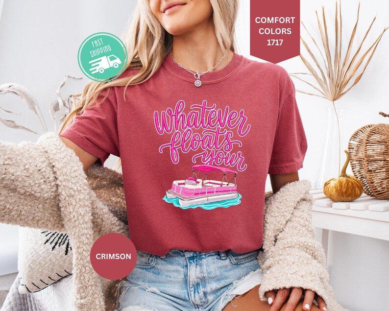 Op de afbeelding: Karmizijnrood T-shirt met de tekst "Whatever floats your" in roze script, met een roze en witte pontonboot afbeelding. Het model draagt een spijkerbroekshort en een cr&egrave;me vest. Een snelle verzendicoon is zichtbaar.
