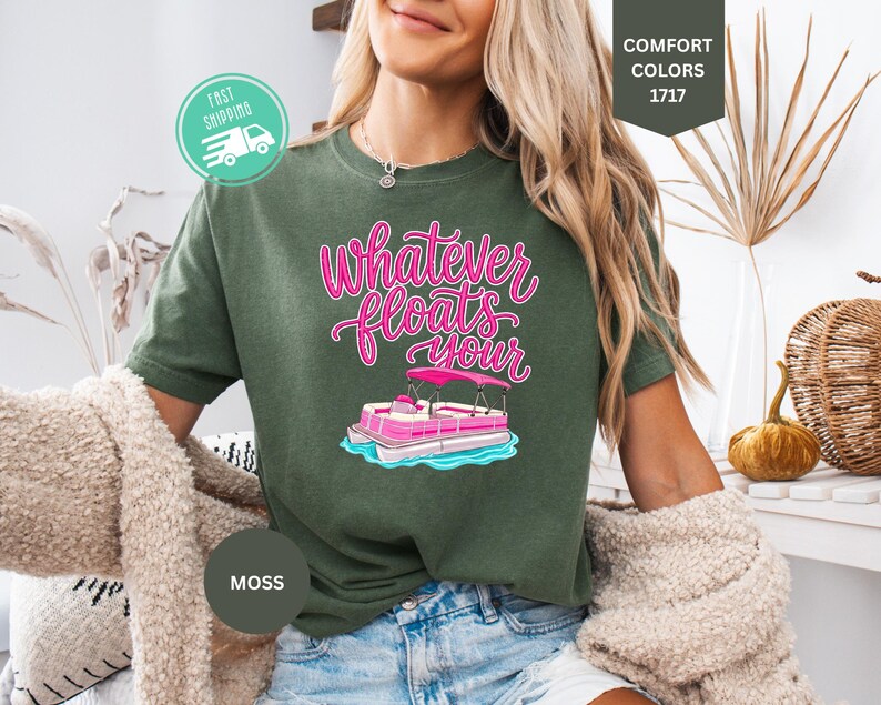 Op de afbeelding: Mosgroen T-shirt met de tekst "Whatever Floats Your" in roze script, met een roze en witte pontonbootafbeelding. Het shirt is van het merk Comfort Colors.