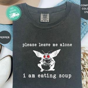 May include: A charcoal gray crew neck t-shirt featuring a white graphic of a creature with large ears and red eyes holding a bowl and spoon. The text above the graphic reads please leave me alone and the text below reads i am eating soup.