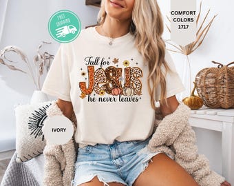 May include: An ivory short-sleeve t-shirt featuring a graphic print with the text Fall for JESUS he never leaves. The lettering is filled with an orange and brown leopard print pattern, accented by small illustrations of pumpkins, leaves, and flowers.